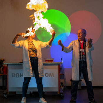 Zwei Personen in weißen Laborkitteln führen eine wissenschaftliche Vorführung durch, bei der eine Flamme über den Händen einer Person aufsteigt, bunte Lichtkreise sind an der Wand zu sehen.