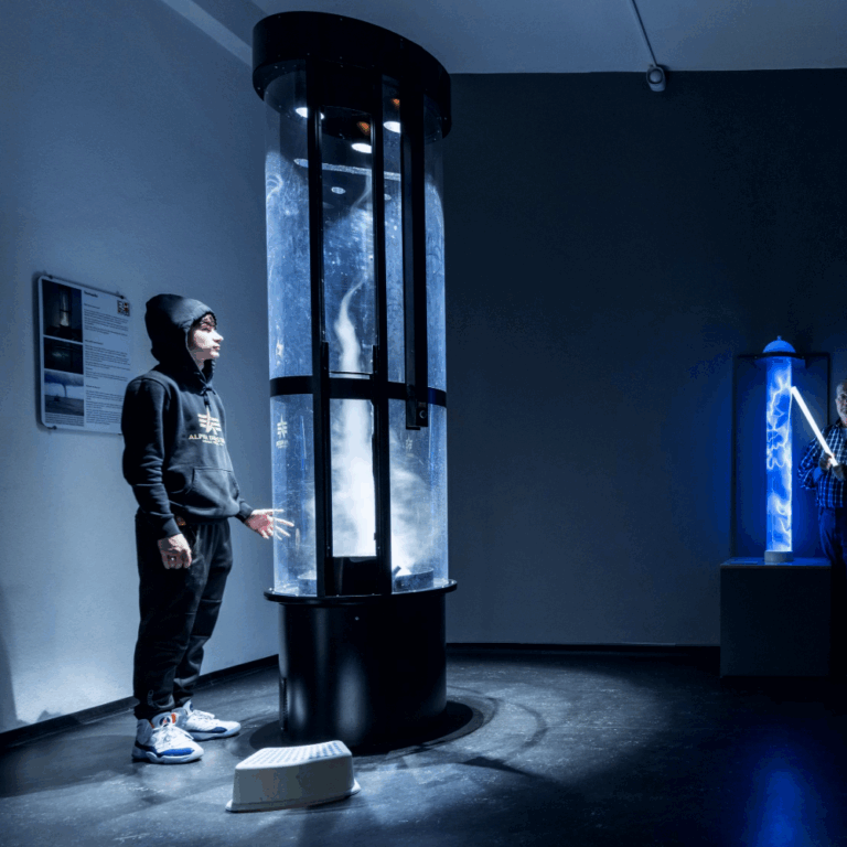 Dunkler Ausstellungsraum mit drei Personen, die sich um zwei beleuchtete, zylindrische Kunstinstallationen bewegen.