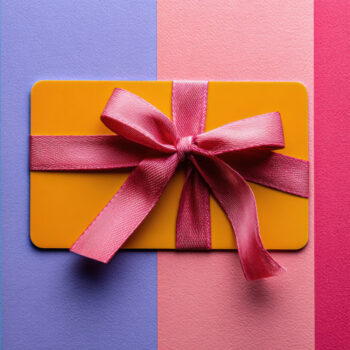 Orangefarbene Karte mit rosa Schleife, liegt auf einem Hintergrund aus drei vertikalen Streifen in Lila, Hellrosa und Dunkelrosa.