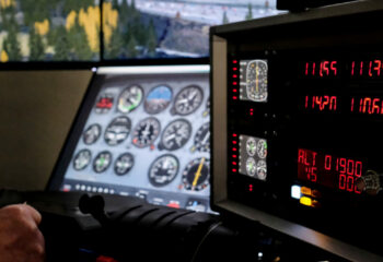 Cockpit eines Flugsimulators mit Instrumententafel, Steuerknüppel und digitaler Anzeige, im Hintergrund Flughafen und Landebahn sichtbar.