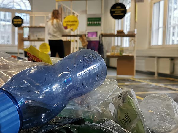 Auf dem Bild sieht man eine blaue Plastikflasche. Im Hintergrund ist die Pop-Up-Ausstellung Plastikfrei denken, nachhaltig handeln zu sehen.