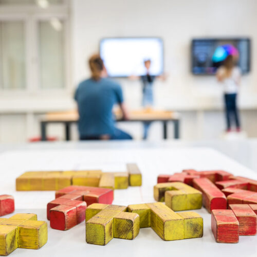 Rote und gelbe Holzbausteine in verschiedenen Formen auf weißer Oberfläche, unscharfer Hintergrund mit Stühlen und Tischen.