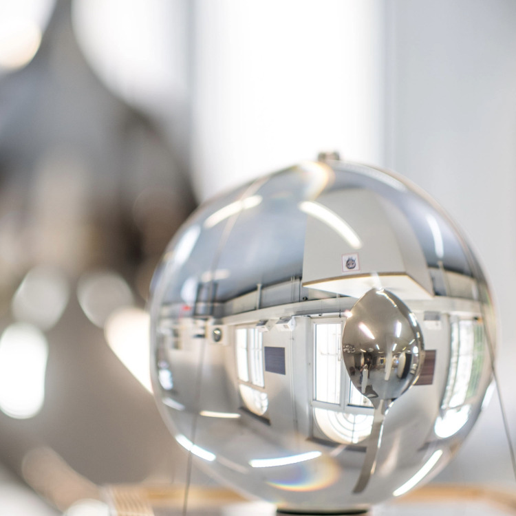 Glaskugel auf Holzsockel in einem hellen Raum mit großen Fenstern und unscharfen Personen im Hintergrund.