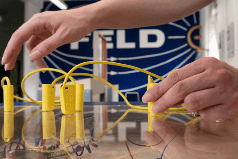 Hände greifen gelbe Kunststoffteile mit Drähten auf einem Tisch, im Hintergrund unscharf das Wort 'FELD' auf blauem Grund.