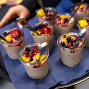 Glas mit Joghurt oder Pudding, garniert mit Beeren und Fruchtstücken, auf blauem Tuch auf einem Tablett.