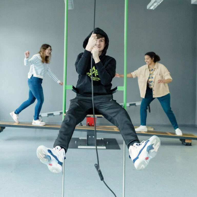 Person in schwarzer Kleidung sitzt auf Schaukel mit gespreizten Beinen, zwei weitere Personen stehen auf Holzbrett im Hintergrund.