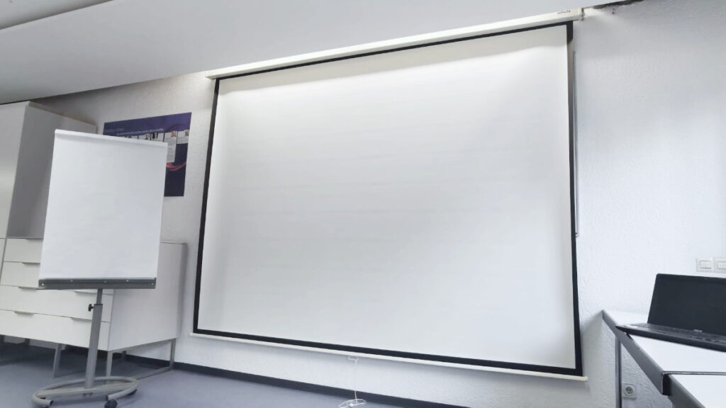 Leinwand mit weißer Projektionsfläche in einem Raum, links ein Flipchart, rechts ein Laptop auf einem Tisch.