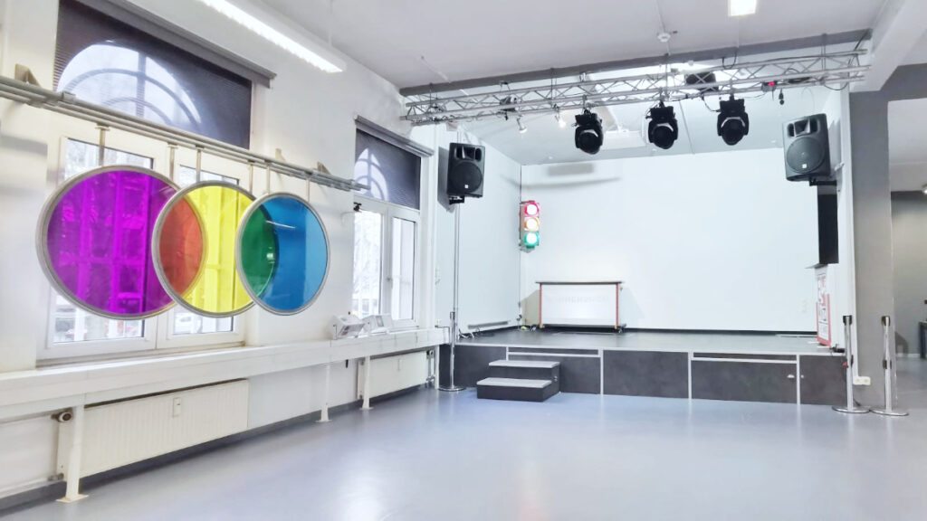 Leerer heller Raum mit Bühne, Treppenstufen und drei farbigen, runden Scheiben an der Fensterfront.