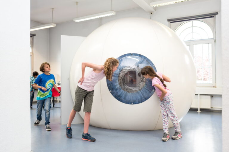 Zwei Mädchen blicken neugierig in ein großes, kugelförmiges Modell eines menschlichen Auges in einem hellen Raum mit hohen Fenstern.