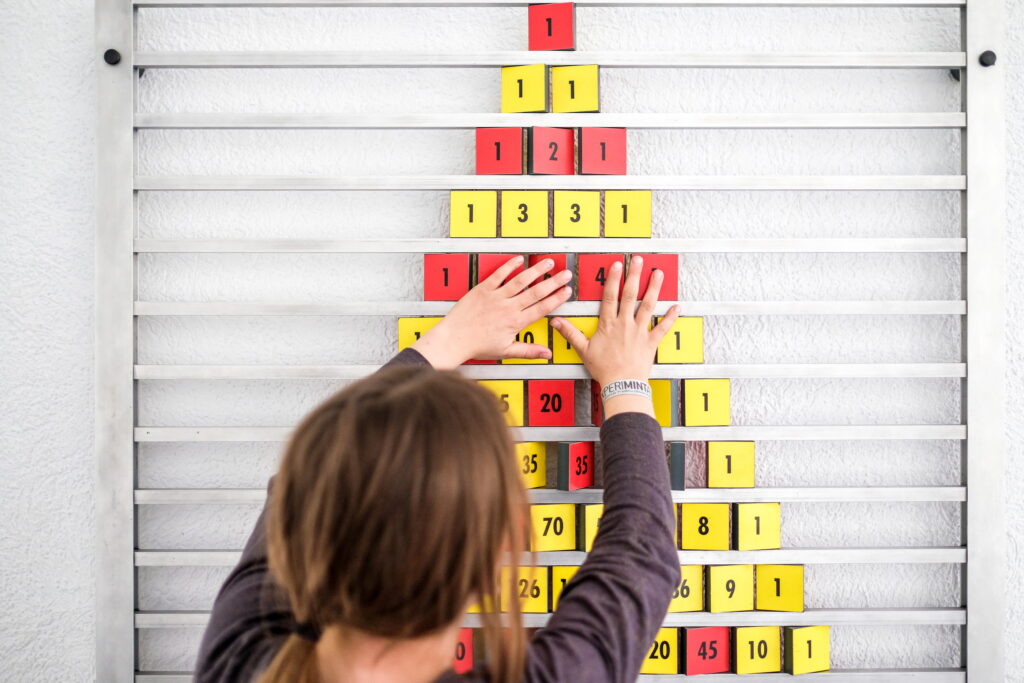 Person von hinten ordnet rote und gelbe Zahlenplättchen in Pyramidenform an einer weißen Wand mit horizontalen Leisten.