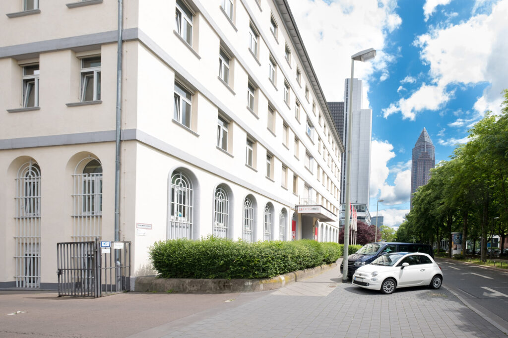 Straßenszene mit Eingang zum Experimenta Science Center, grüner Hecke und Hochhaus im Hintergrund unter blauem Himmel mit Wolken.
