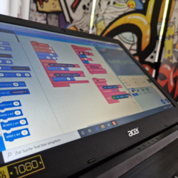 Laptopbildschirm zeigt eine Programmieroberfläche mit farbigen Codeblöcken in Blau und Pink, darunter Suchleiste mit Text 'Zur Suche Text hier eingeben'.