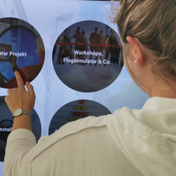 Person mit weißem Pullover und Uhr berührt interaktiven Bildschirm mit Kreisen, Texten wie 'Workshops, Flugsimulator & Co.' sichtbar.