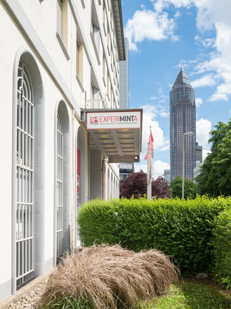 Straßenszene mit Eingang zum Experimenta Science Center, grüner Hecke und Hochhaus im Hintergrund unter blauem Himmel mit Wolken.
