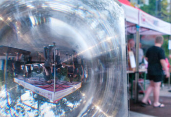 Vergrößernde Linse zeigt auf einem Marktstand eine umgekehrte Spiegelung von Personen und einem Zelt mit rotem Dach.