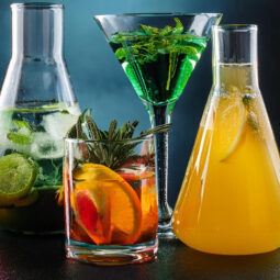 Vier Gläser mit verschiedenen Getränken: ein Glas mit Limettenscheiben, ein Glas mit Orangenscheiben und Rosmarin, ein Cocktailglas mit grünen Kräutern, ein Kolben mit gelbem Saft und Zitronenscheiben.