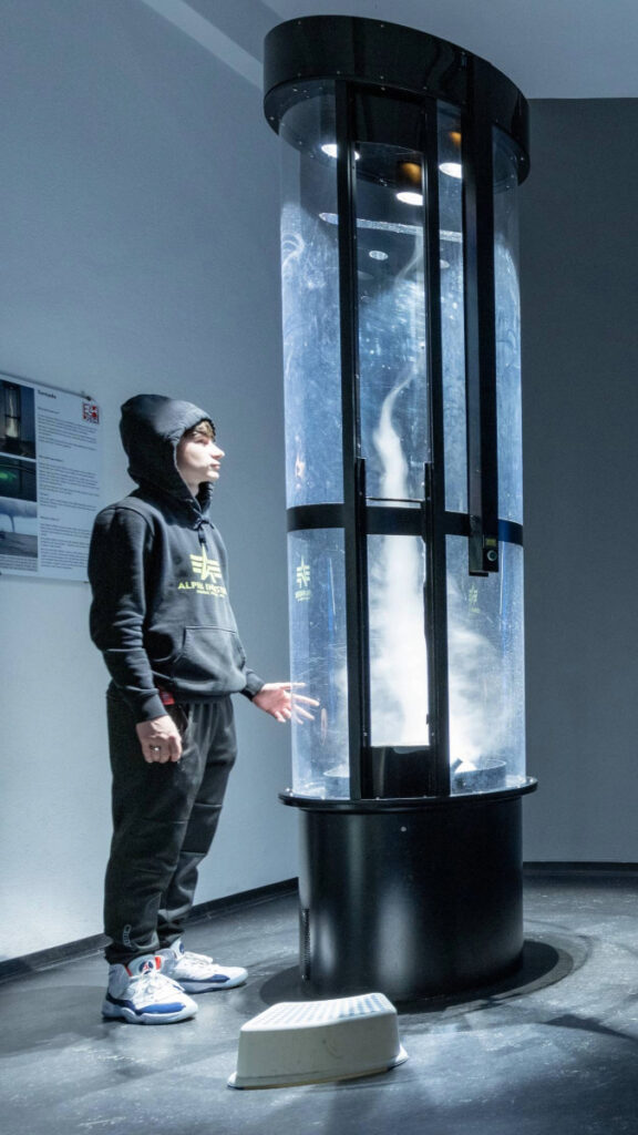 Dunkler Ausstellungsraum mit einer Person, die sich um eine beleuchtete, zylindrische Kunstinstallationen bewegt.