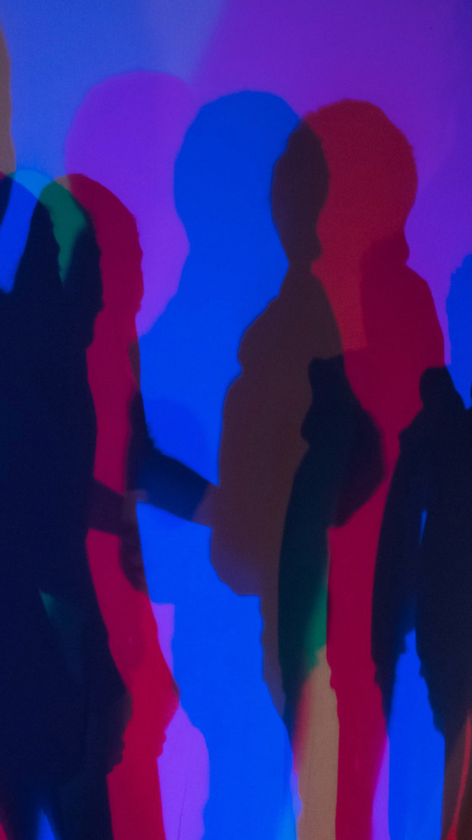 Mehrere farbige Schatten von Personen auf einer Wand, überwiegend in Blau, Rot und Grün.