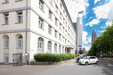 Straßenszene mit Eingang zum Experimenta Science Center, grüner Hecke und Hochhaus im Hintergrund unter blauem Himmel mit Wolken.