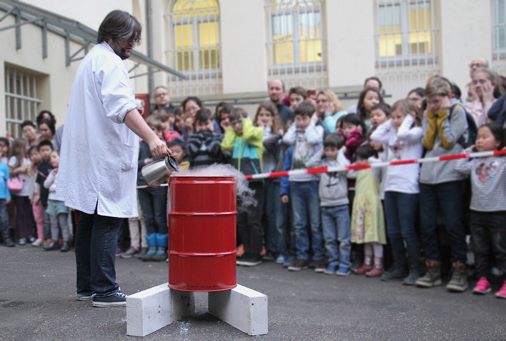 Person in weißem Kittel gießt Flüssigkeit in ein rotes Fass auf einem Betonständer, umgeben von einer Gruppe zuschauender Kinder und Erwachsener hinter Absperrband.