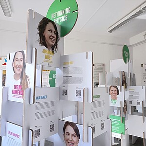Mehrere weiße Ausstellungstafeln mit Texten und QR-Codes, darüber grüne Schilder mit der Aufschrift 'RETHINKING PHYSICS' in einem hellen Raum.