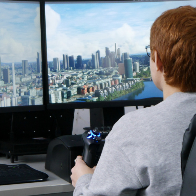 Person mit rotem Haar sitzt vor zwei großen Bildschirmen und hält einen Joystick, auf den Bildschirmen ist eine Stadt mit Hochhäusern und Fluss zu sehen.