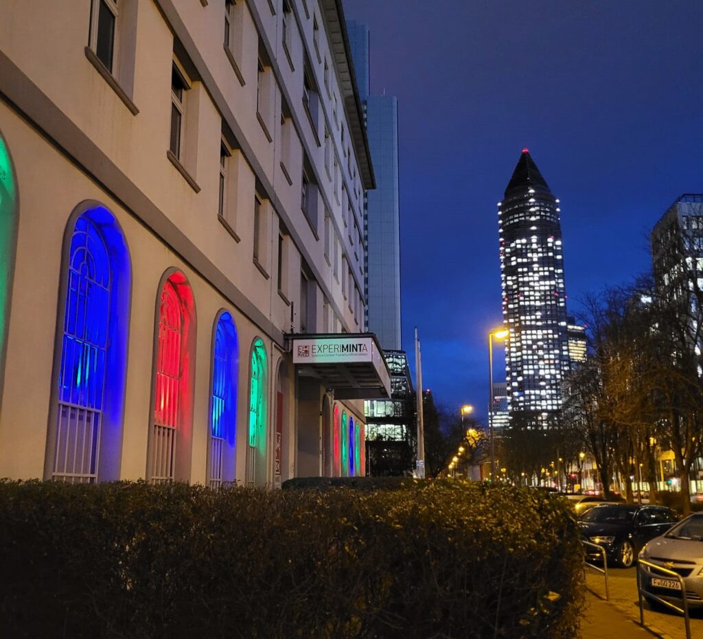 Gebäudefassade mit bunten, blau, rot und grün beleuchteten Fenstern, daneben Straßenszene bei Nacht mit beleuchtetem, spitz zulaufendem Wolkenkratzer im Hintergrund.