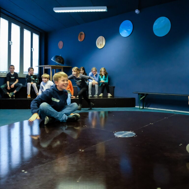 Mehrere Kinder sitzen und stehen in einem Raum mit dunkelblauen Wänden, auf denen bunte runde Dekorationen hängen und einer großen runden Scheibe die sich drehen kann
