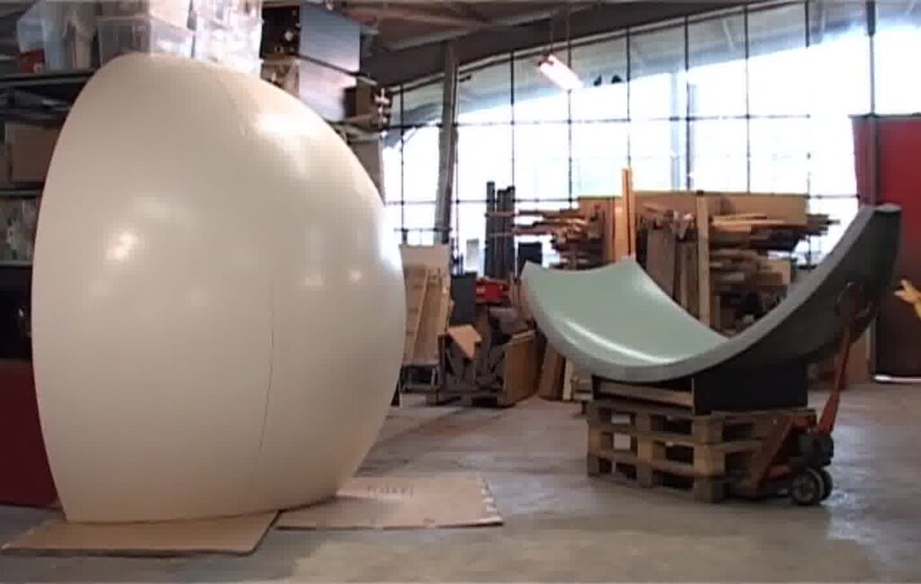 Großer, weißer, runder Gegenstand links und zwei gebogene, glatte Objekte auf Holzpaletten rechts in einer Werkhalle mit Holzstapeln und großen Fenstern.